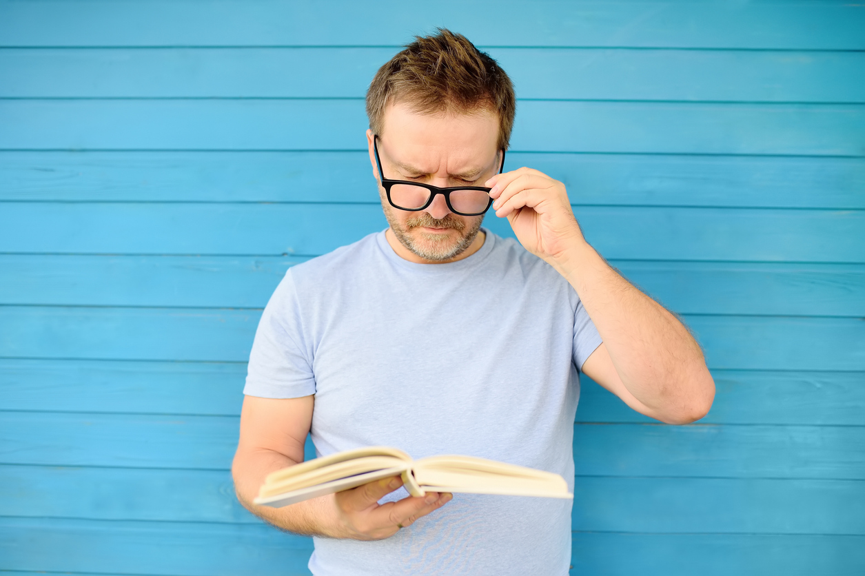 Portrait of mature man with big black eye glasses trying to read book but having difficulties seeing text because of vision problems
